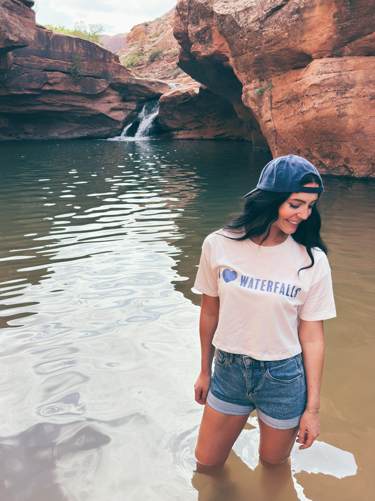 Woman standing in water near rocky cliffs with a t-shirt that says 'I Love Waterfalls'.