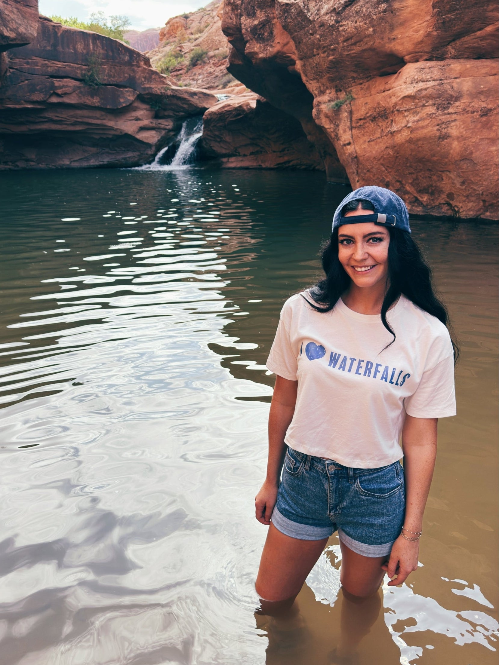 Woman standing in a natural rock formation with water, wearing a 'I love Waterfalls' t-shirt.